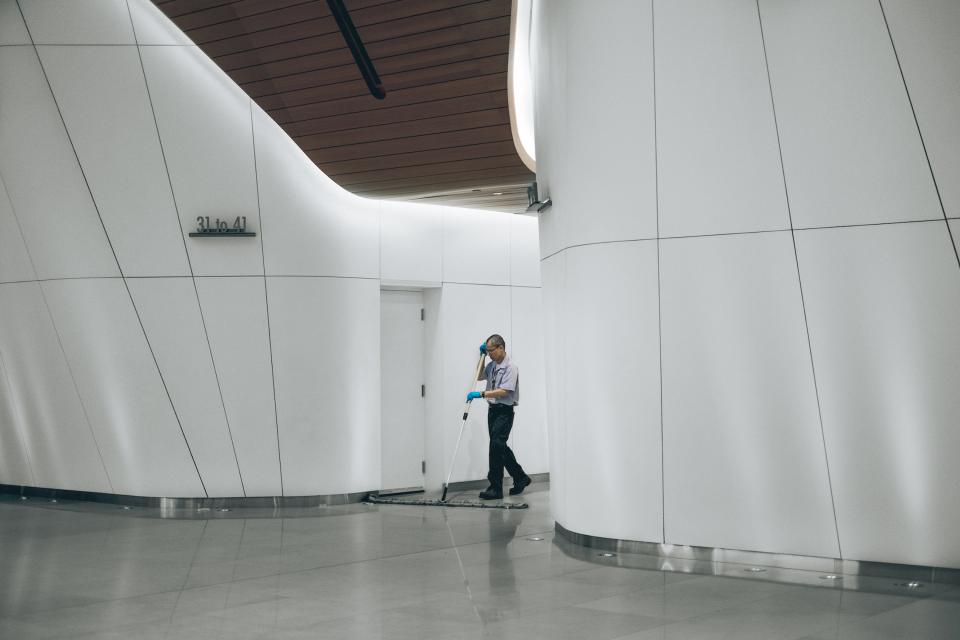 Een schoonmaakmedewerker in uniform en handschoenen veegt een glanzende vloer in een moderne, witte bedrijfsruimte met g